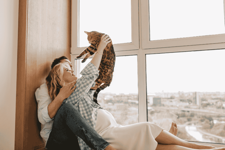 Casal sentado à beira da janela segurando seu gato, representando afeto, planejamento e compromisso a longo prazo com o pet.