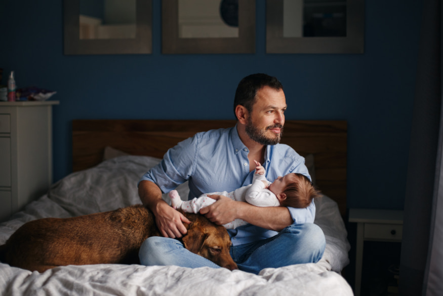 Pai segurando bebê recém-nascido no colo enquanto acaricia o cão deitado na cama, ilustrando a adaptação do cachorro à chegada do novo bebê em casa.