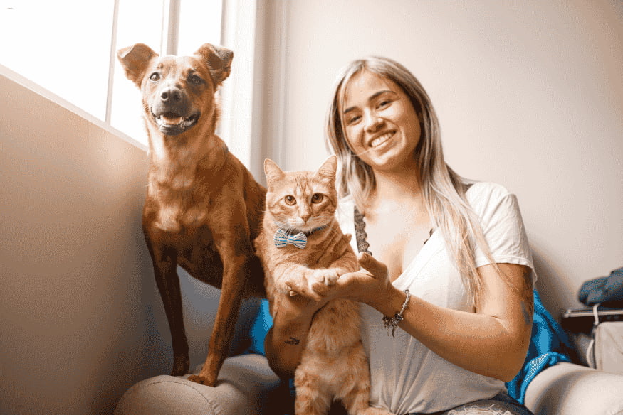 Mulher sorridente segurando um gato com laço no colo ao lado de um cachorro.