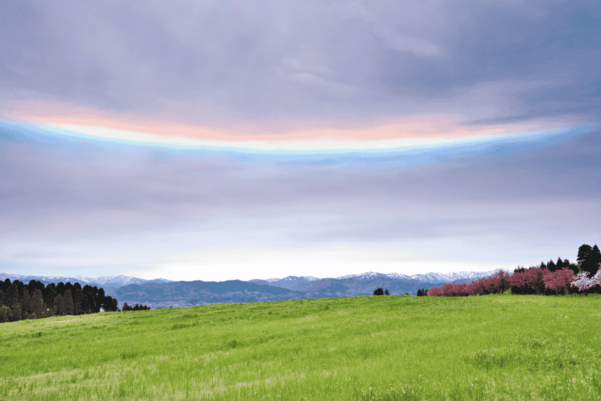 Arco circun-horizontal colorido atravessando o céu acima de campo verde e montanhas ao fundo, com aparência de arco-íris invertido.
