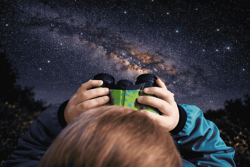 Pessoa usando binóculo para observar o céu noturno, com a Via Láctea visível ao fundo em ambiente escuro.