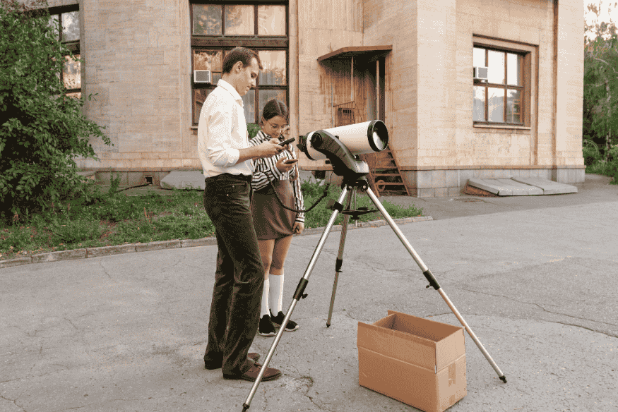 Adulto e criança montando e ajustando telescópio em área externa, mostrando educação científica e participação popular na astronomia.