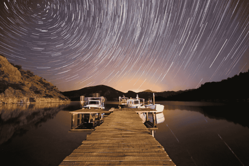 Píer sobre lago com barcos parados sob céu cheio de trilhas de estrelas, em fotografia noturna de longa exposição.