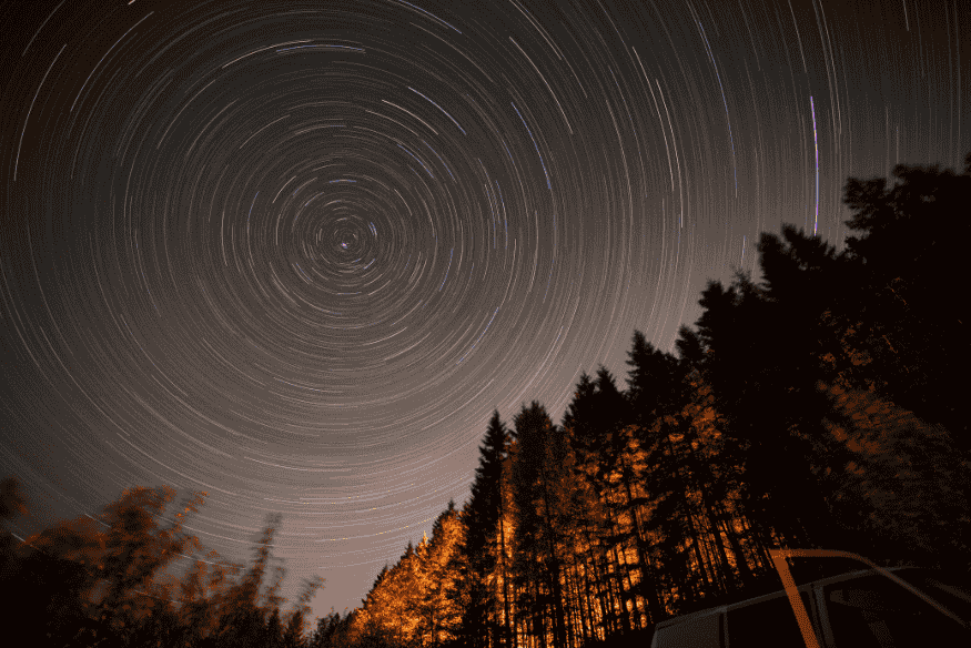 Trilhas de estrelas circulares no céu noturno sobre floresta escura, com efeito de rotação ao redor do polo celeste em longa exposição.