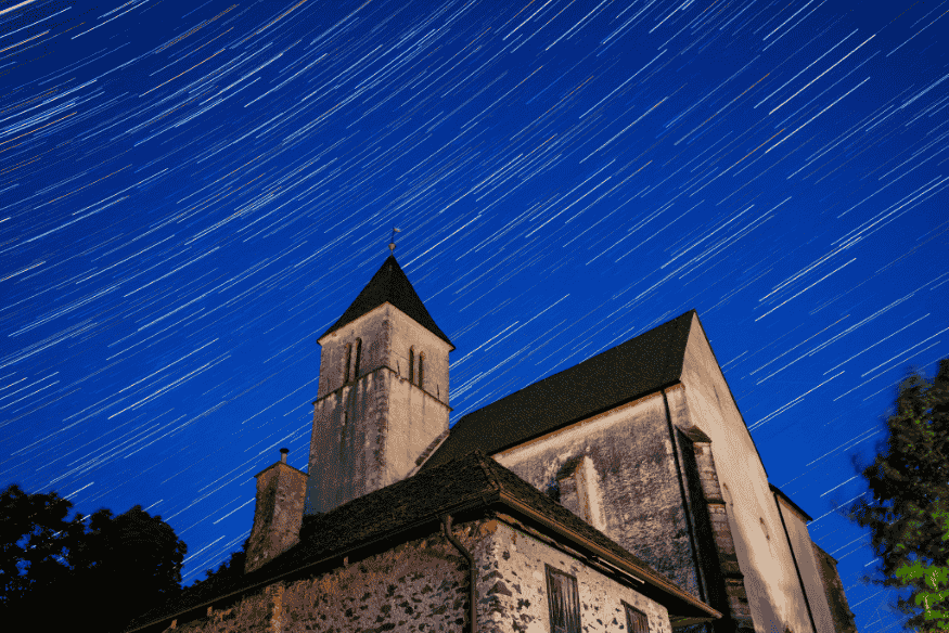 Igreja iluminada sob céu noturno com trilhas de estrelas em longas linhas, exemplo de fotografia star trails com elemento arquitetônico em primeiro plano.