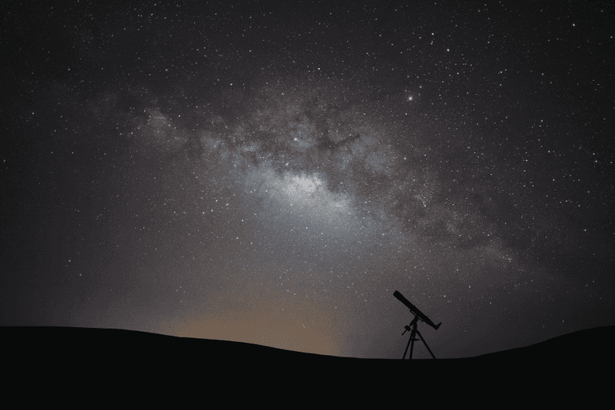 Telescópio em silhueta sob céu noturno com faixa da Via Láctea visível, em paisagem escura ideal para observação astronômica.