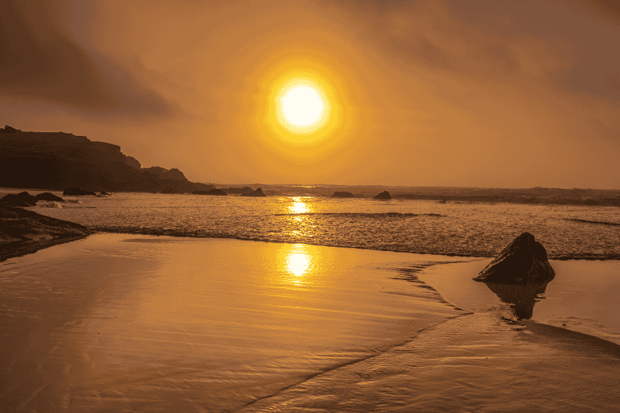 Sol brilhando sobre o mar ao entardecer, com reflexo dourado na água e rochas na praia.