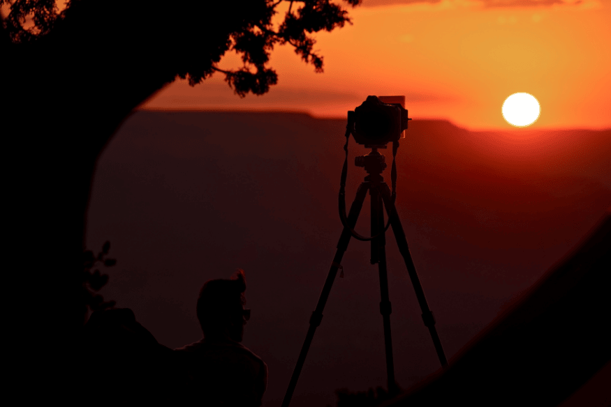 Câmera em tripé voltada para o Sol baixo no horizonte, em silhueta contra céu alaranjado ao entardecer.