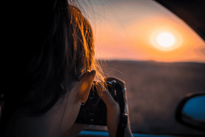 Pessoa fotografando o Sol ao entardecer com câmera na janela de um carro, em cena relacionada à observação do astro.