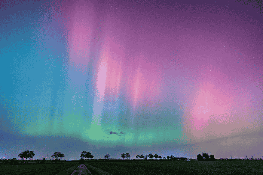 Aurora austral em tons rosa, roxo e verde sobre campo aberto durante a noite, com feixes luminosos no céu.