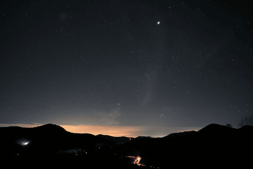 Paisagem montanhosa sob céu noturno com vários pontos luminosos, mostrando a observação de estrelas e possíveis planetas visíveis sem telescópio.