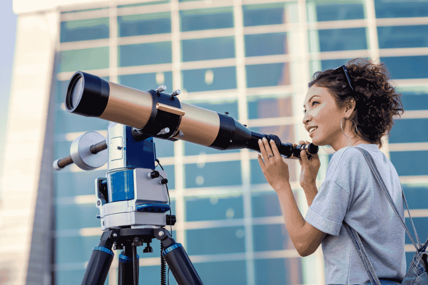 Mulher observando pelo telescópio refrator montado em tripé, em ambiente urbano ao ar livre durante o dia.