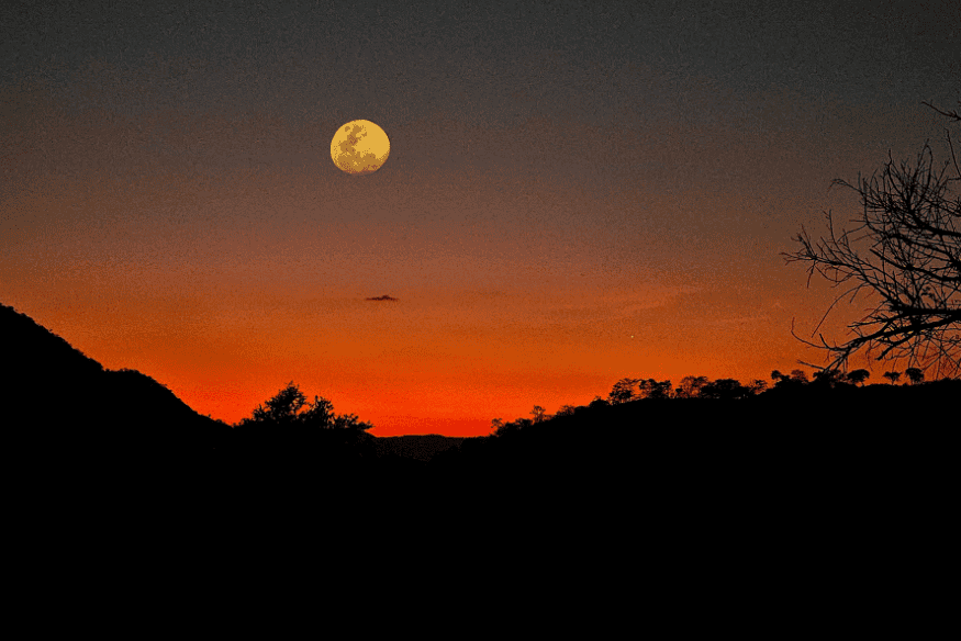 Lua cheia acima de horizonte alaranjado ao entardecer, com silhuetas de morros e árvores compondo a paisagem.