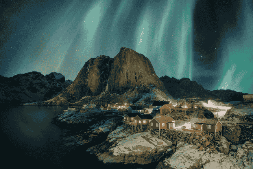 Aurora boreal intensa sobre vila costeira e montanhas nevadas, com faixas luminosas verdes no céu refletidas sobre a paisagem noturna.