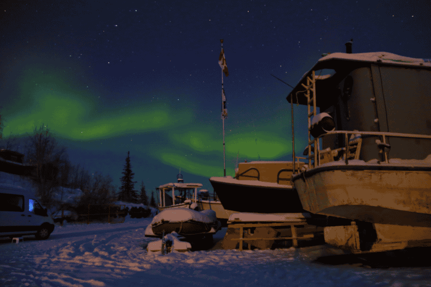 Aurora boreal verde no céu noturno sobre barcos cobertos de neve, em cena de inverno com estrelas visíveis ao fundo.