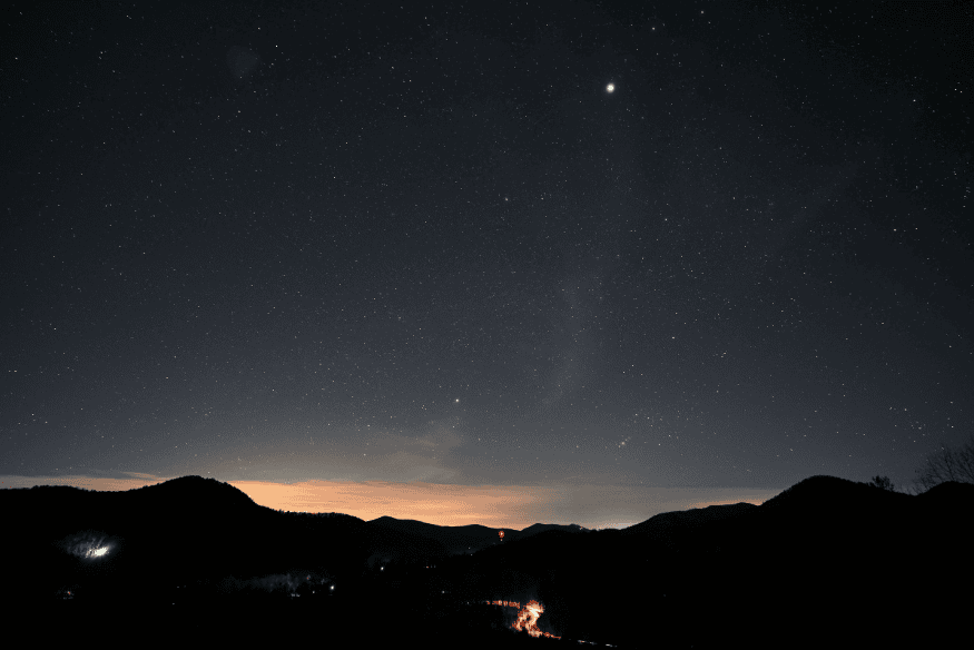 Céu noturno estrelado sobre montanhas escuras, com horizonte iluminado ao fundo em paisagem ampla ideal para fotografia com celular.