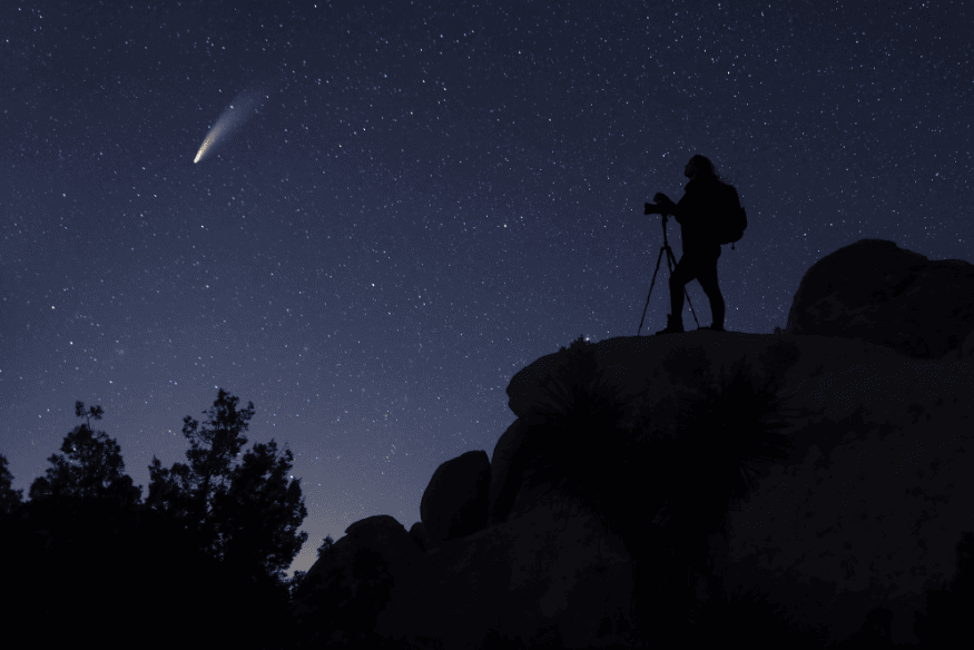 Pessoa fotografando o céu noturno com câmera em tripé sobre rochas, enquanto um cometa aparece brilhante entre as estrelas.