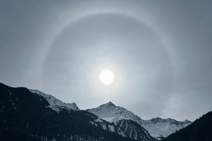 Halo solar sobre montanhas cobertas de neve, com grande anel luminoso ao redor do Sol em céu nublado.