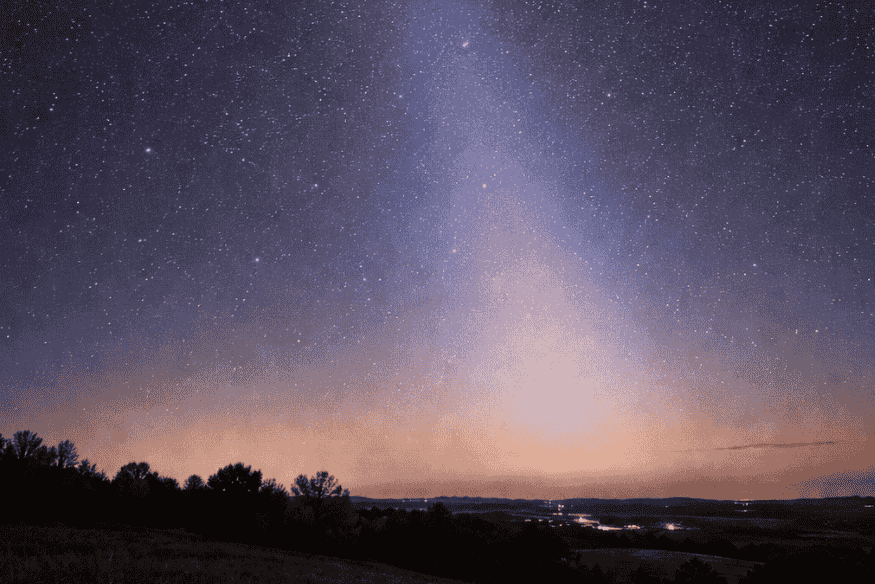 Luz zodiacal visível como um cone de brilho suave subindo do horizonte em céu estrelado, sobre paisagem escura com árvores e luzes distantes.