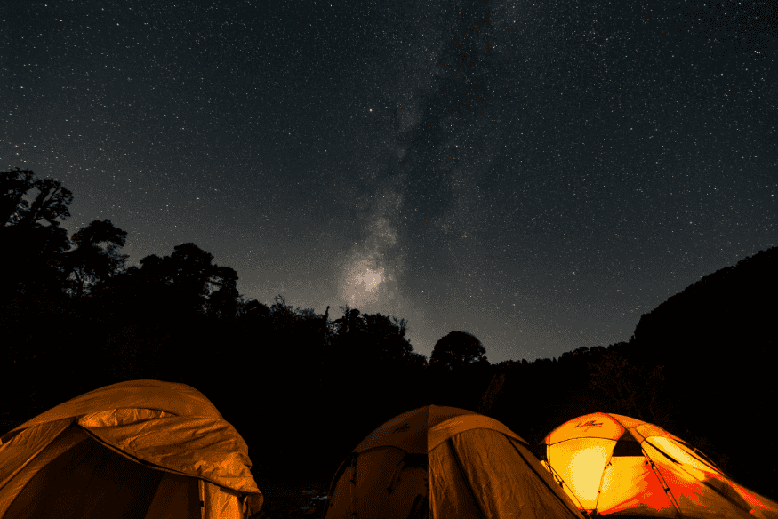 Barracas iluminadas em área de camping sob céu estrelado com a Via Láctea visível, em local escuro ideal para observação astronômica.