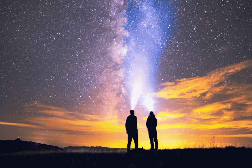 Duas pessoas observando o céu noturno em campo aberto, com a Via Láctea visível acima do horizonte e pouca luz artificial ao redor.