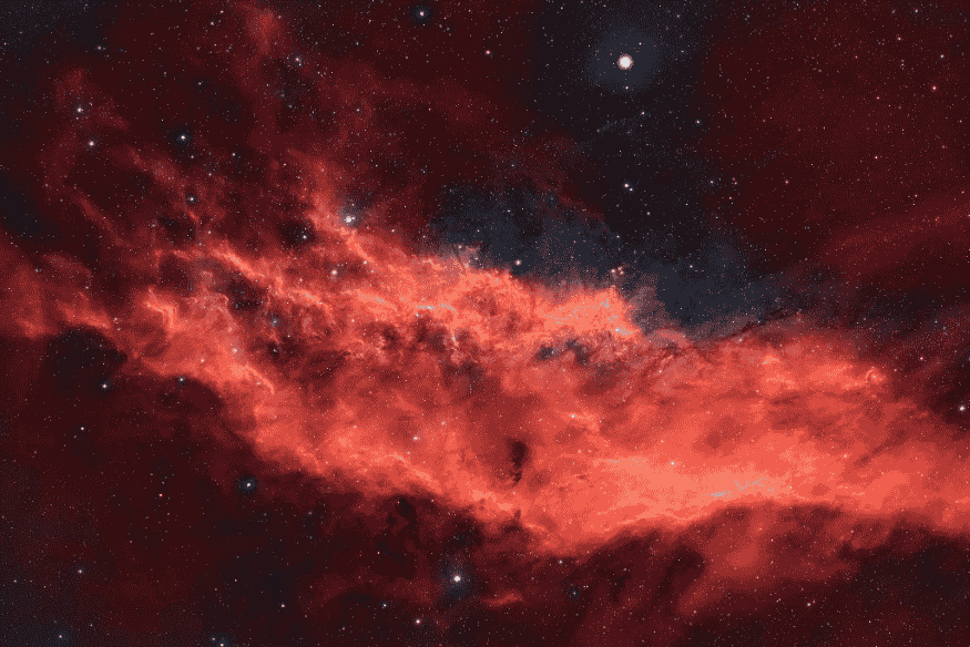 Nebulosa avermelhada com filamentos de gás brilhante espalhados pelo espaço, destacando sua estrutura extensa entre estrelas.