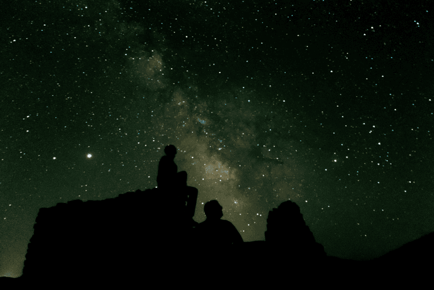 Silhuetas de pessoas observando o céu estrelado a olho nu, com a Via Láctea visível ao fundo em uma noite escura.