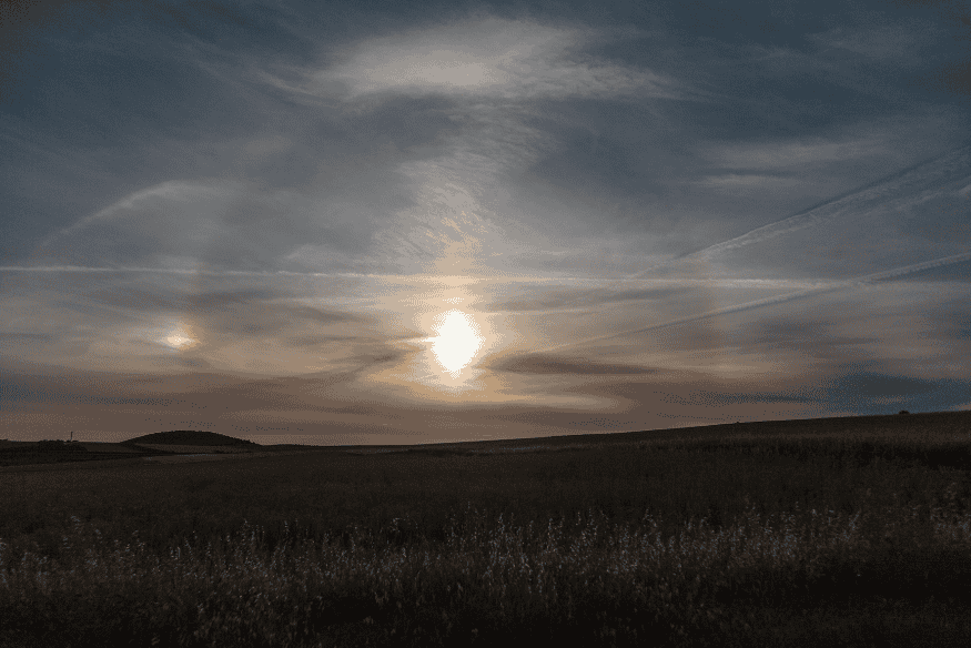 Parélio em céu com nuvens finas sobre campo aberto, mostrando brilho lateral ao Sol causado por cristais de gelo na atmosfera.