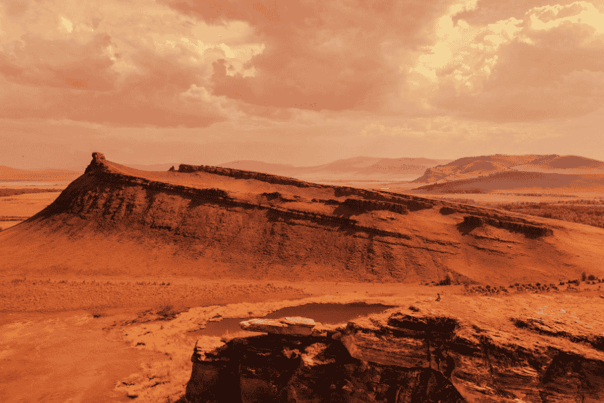 Paisagem de Marte com terreno rochoso, montanhas e céu alaranjado, representando o ambiente seco do planeta.