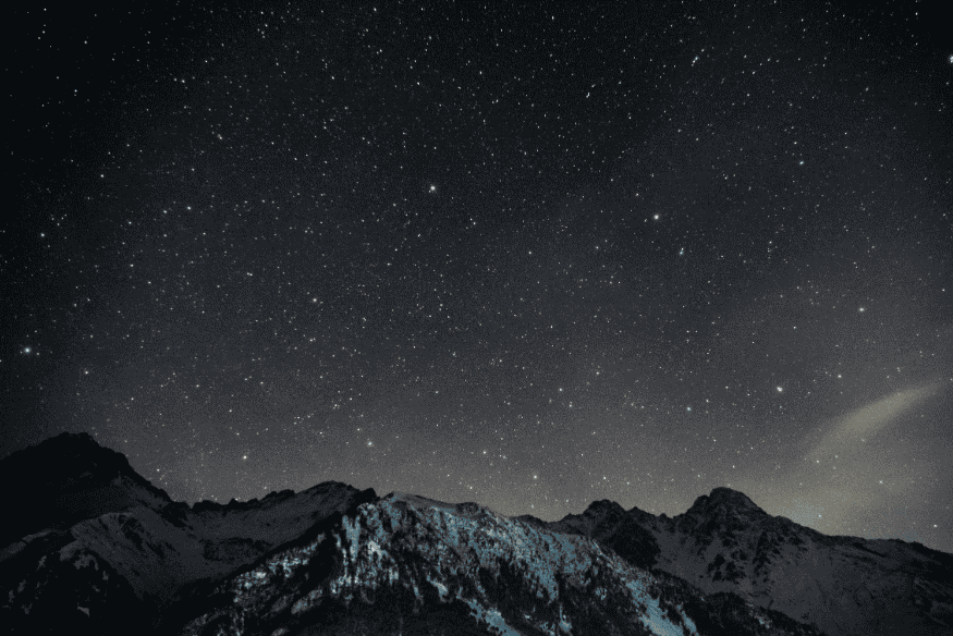 Paisagem montanhosa sob céu estrelado escuro, mostrando como o espaço entre as estrelas domina a aparência do céu noturno visto da Terra.