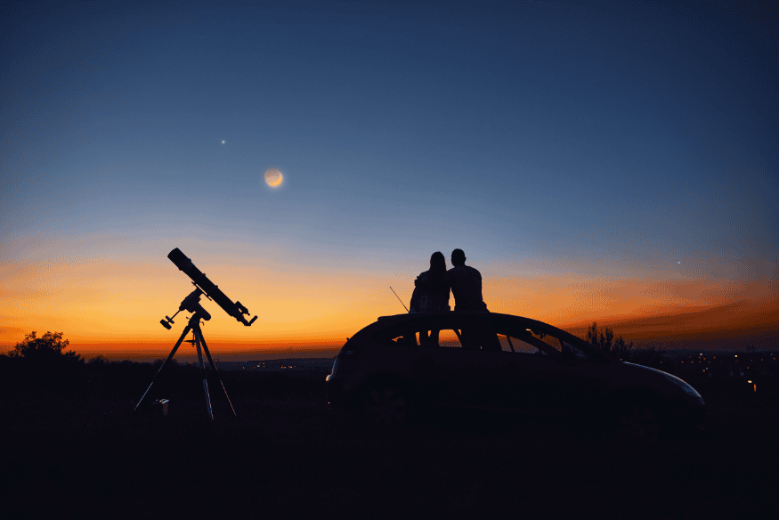 Casal ao lado de um carro e de um telescópio ao entardecer, observando o céu em uma sessão de astronomia ao ar livre.