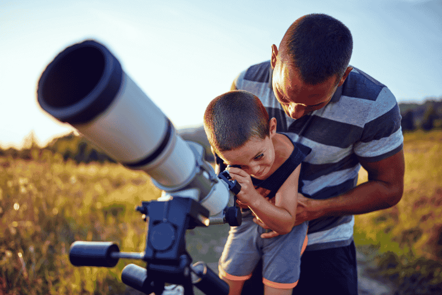 Adulto ajudando uma criança a usar um telescópio ao ar livre, em cena que representa uma sessão de observação do céu em família.