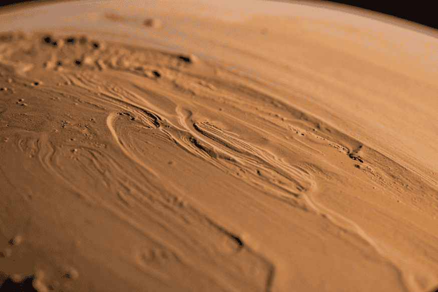Close da superfície de Marte com relevo marcado por sulcos, camadas de poeira e formações onduladas em tons alaranjados.